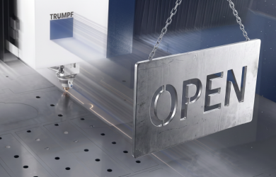 Pravokotna kovinska tablica z lasersko izrezanim napisom "OPEN", ki visi na kovinski verigi pred delujočim fiber laserjem Trumpf 12kW med razrezom pločevine.